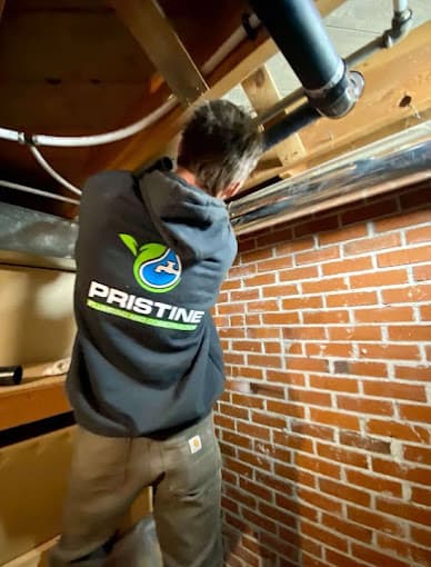 A Pristine Plumbing technician in a branded hoodie working on overhead pipework in a basement